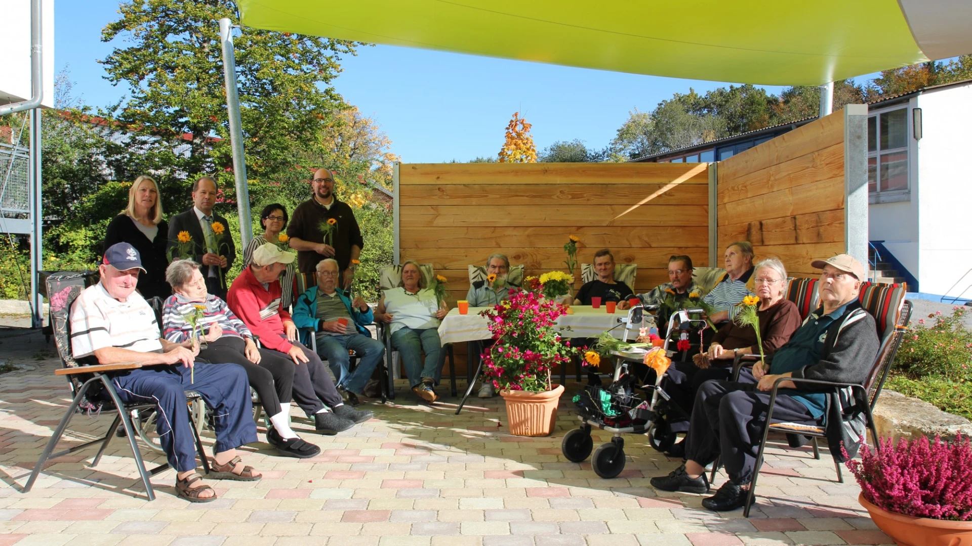 Eine Gruppe von Menschen, die unter einen gelben Sonnensegel sitzen. Sie befinden sich auf einer gepflasterten Fl&auml;che mit Holzwand im Hintergrund. Vor ihnen stehen mehrere mit Blumen bepflanzte T&ouml;pfe und Schalen und ein Tisch mit Bechern.