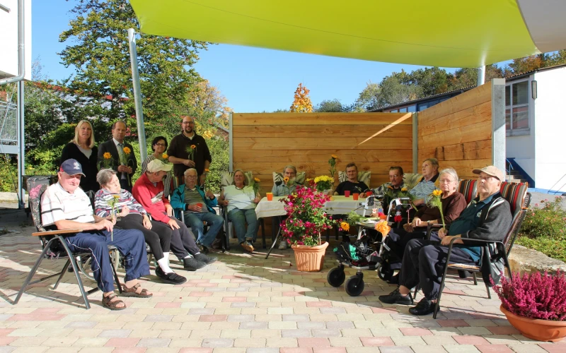 Eine Gruppe von Menschen, die unter einen gelben Sonnensegel sitzen. Sie befinden sich auf einer gepflasterten Fl&auml;che mit Holzwand im Hintergrund. Vor ihnen stehen mehrere mit Blumen bepflanzte T&ouml;pfe und Schalen und ein Tisch mit Bechern.