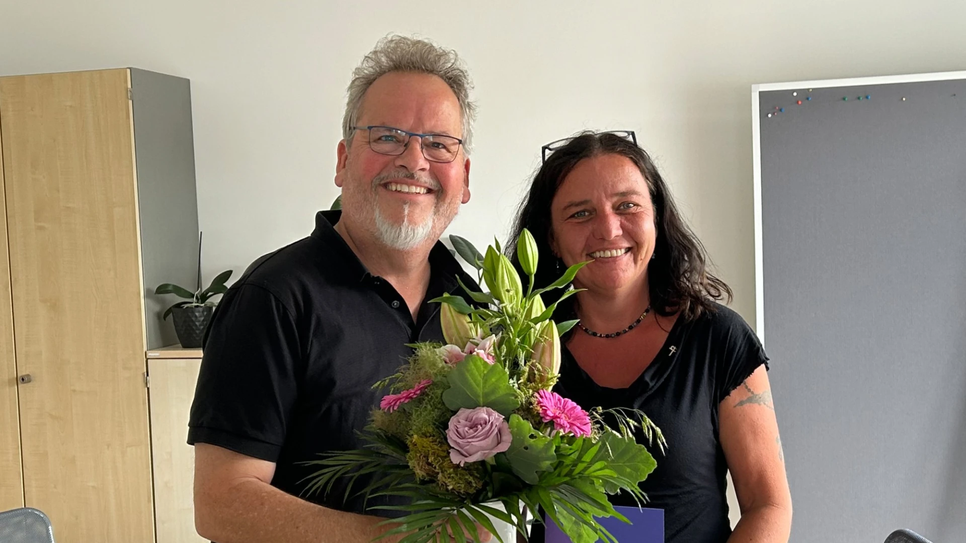 Zwei Personen stehen in einem Besprechungsraum mit Tisch, St&uuml;hlen und einem Schrank und l&auml;cheln in die Kamera. Der Mann links h&auml;lt einen Blumenstrau&szlig; in der Hand und die Frau rechts ein Dokument.