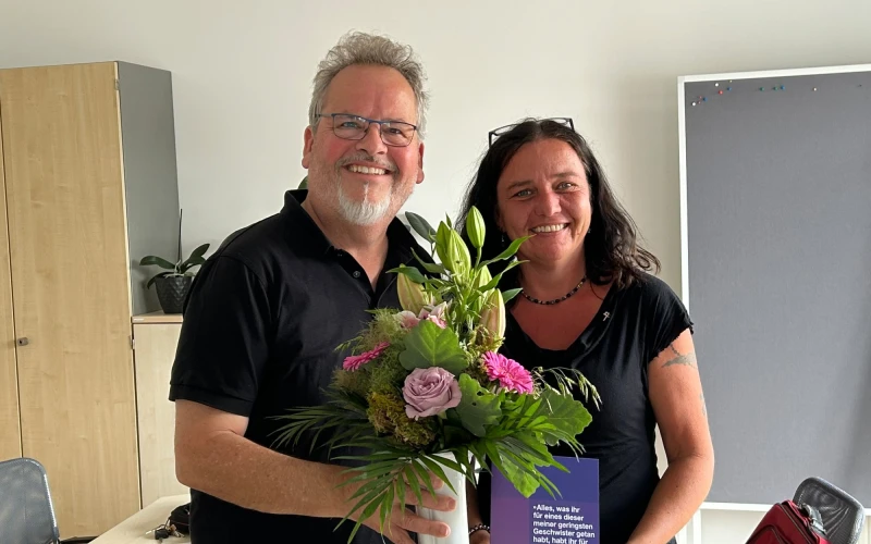 Zwei Personen stehen in einem Besprechungsraum mit Tisch, St&uuml;hlen und einem Schrank und l&auml;cheln in die Kamera. Der Mann links h&auml;lt einen Blumenstrau&szlig; in der Hand und die Frau rechts ein Dokument.