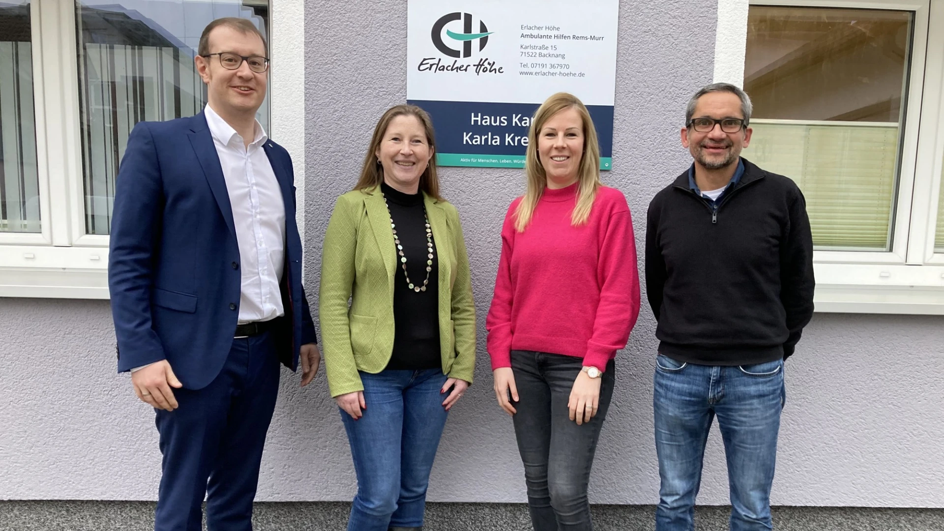 Zwei Frauen stehen zwischen zwei M&auml;nnern an einem Geb&auml;ude. Alle blicken in die Kamera und l&auml;cheln. Im Hintergrund ist ein Schild mit Aufschrift "Erlacher H&ouml;he - Haus Karla" zu sehen.