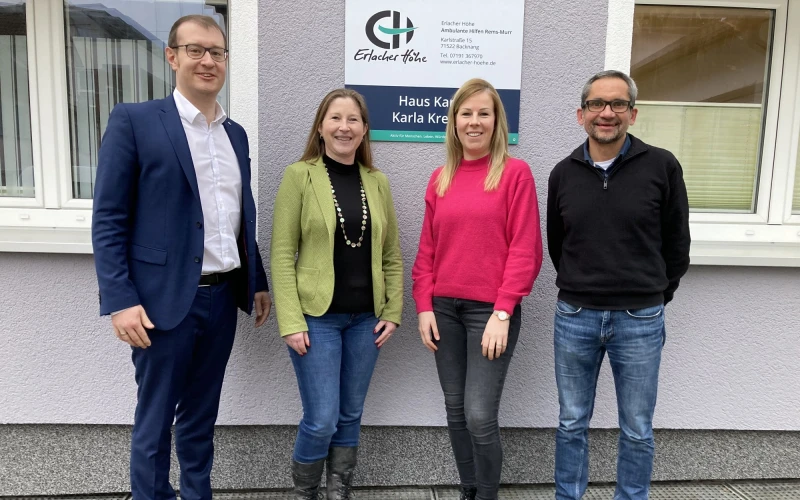 Zwei Frauen stehen zwischen zwei M&auml;nnern an einem Geb&auml;ude. Alle blicken in die Kamera und l&auml;cheln. Im Hintergrund ist ein Schild mit Aufschrift "Erlacher H&ouml;he - Haus Karla" zu sehen.