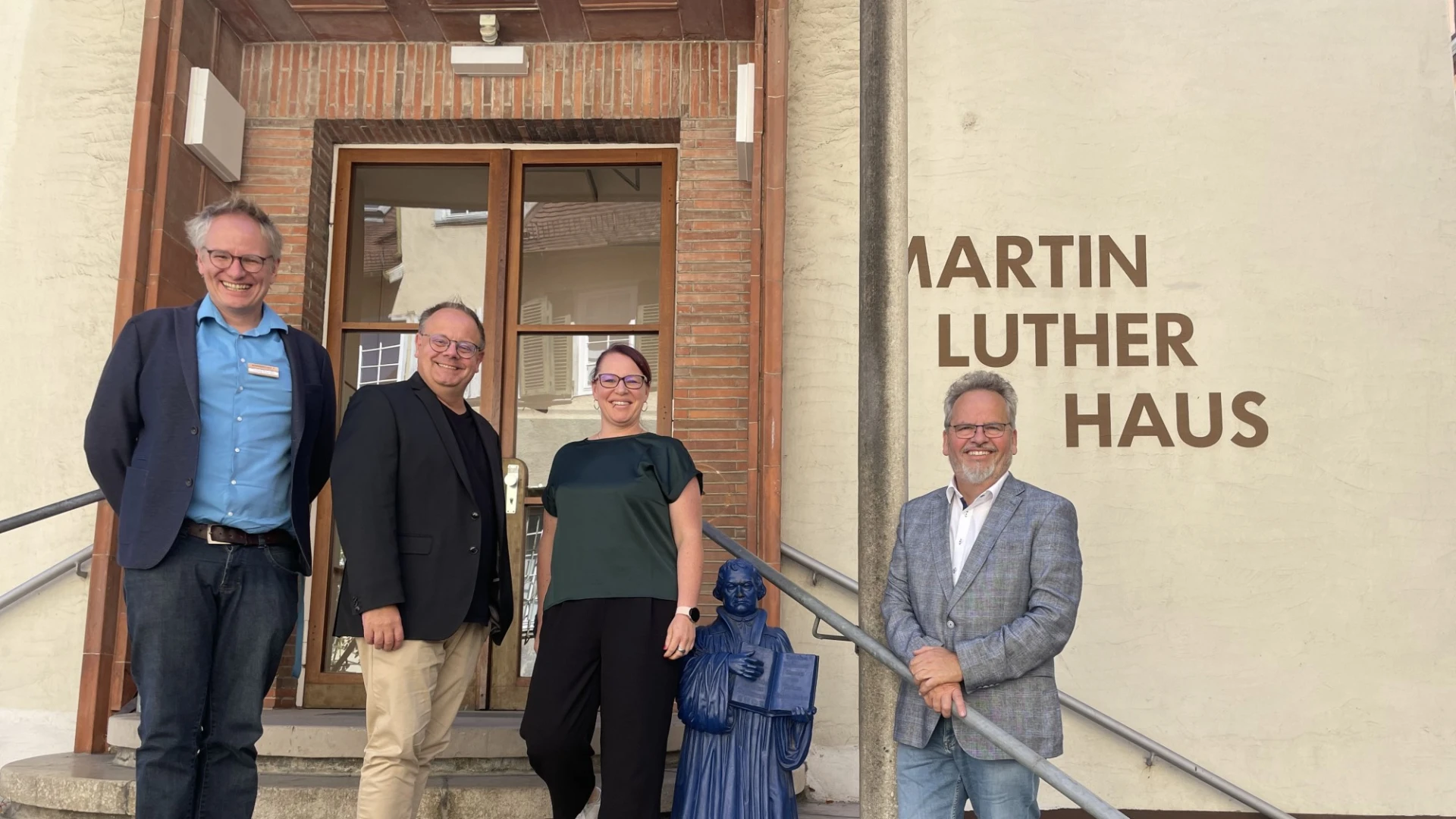 Vier Personen stehen auf den Stufen des Martin-Luther-Hauses vor einer h&ouml;lzernen Eingangst&uuml;r. Auf der Treppe ist eine kleine blaue Figur mit ge&ouml;ffnetem Buch zu sehen.