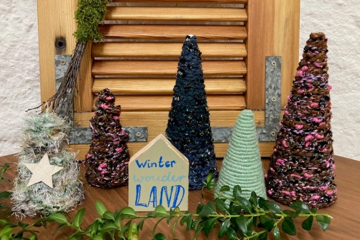 Winterliche Deko-Szene mit verschiedenfarbigen Tannenb&auml;umen mit Garn umwickelt, einem Stern und einem handgeschriebenen Schild "WinterwonderLAND" vor einem Holzjalousie-Hintergrund und wei&szlig;er Wand, dekoriert mit Moos und Zweigen.