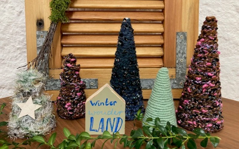 Winterliche Deko-Szene mit verschiedenfarbigen Tannenb&auml;umen mit Garn umwickelt, einem Stern und einem handgeschriebenen Schild "WinterwonderLAND" vor einem Holzjalousie-Hintergrund und wei&szlig;er Wand, dekoriert mit Moos und Zweigen.