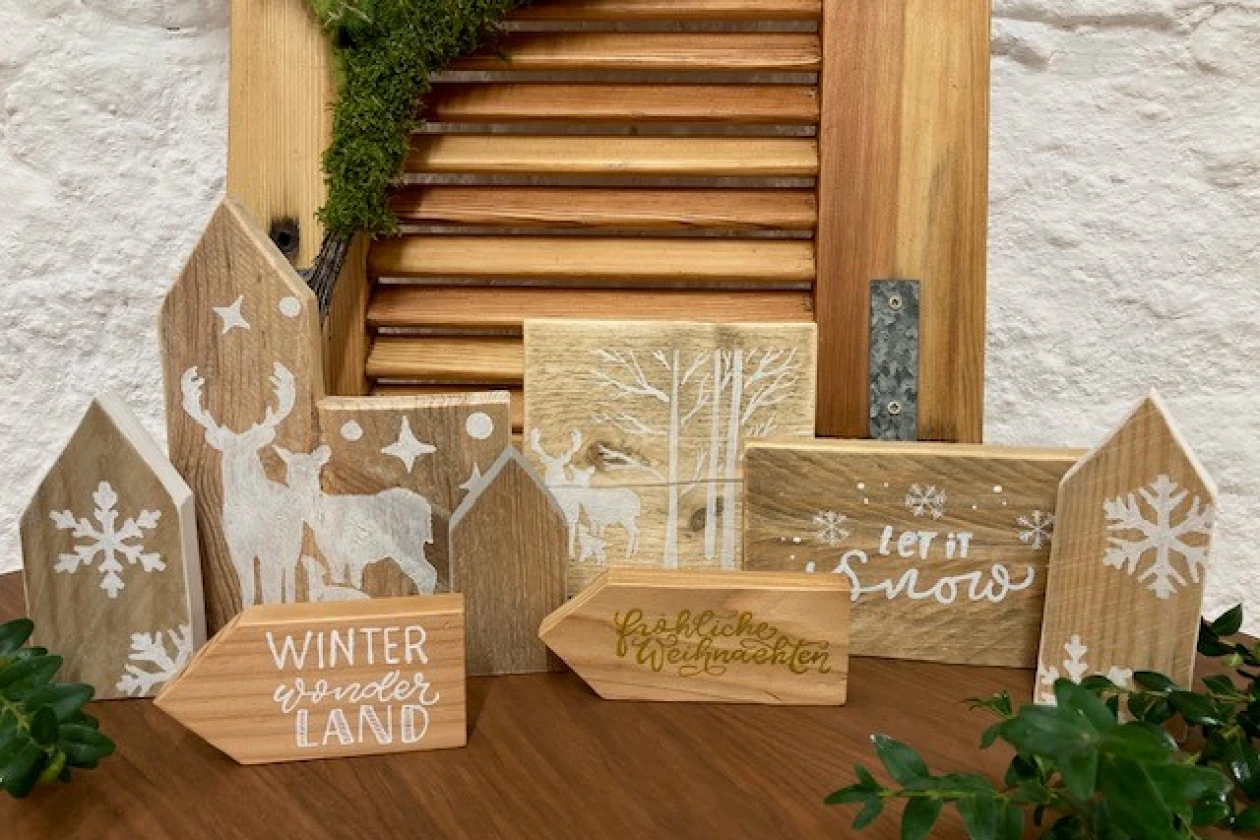 Winterliche Holzdekoration mit verschiedenen Holzh&auml;usern und Schildern, verziert mit Schneeflocken, Rentieren und Wintermotiven, vor einem Holzjalousie-Hintergrund und wei&szlig;er Wand platziert.