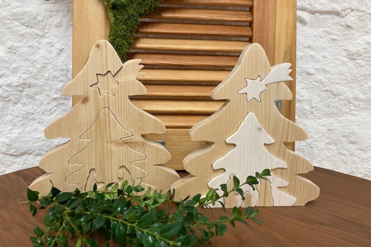 Zwei geschnitzte Holztannen in verschiedenen Gr&ouml;&szlig;en stehen vor einem h&ouml;lzernen Jalousie-Hintergrund, dekoriert mit einem gr&uuml;nen Zweig. Die Tannen haben eine nat&uuml;rliche, helle Holzmaserung und unterschiedliche Sternformen an der Spitze.