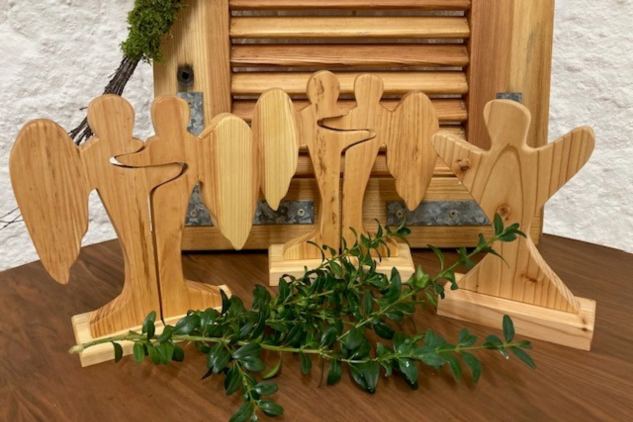 Holzfiguren in Engelsform, aus hellem Holz gefertigt, stehen vor einem Holzjalousie und sind mit gr&uuml;nen Zweigen dekoriert. Die Figuren haben eine stilisierte, abstrakte Form und sind auf einem Holztisch arrangiert.