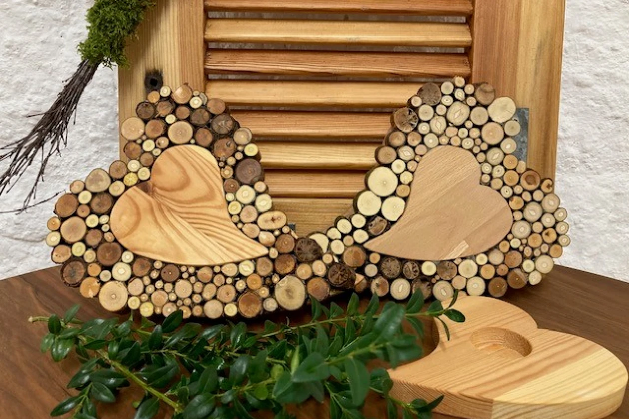 Zwei herzf&ouml;rmige Holzherzen umgeben von kleinen Holzscheiben in verschiedenen Gr&ouml;&szlig;en und Farbt&ouml;nen, vor einem Jalousien-Hintergrund, dekoriert mit gr&uuml;nen Zweigen und einem Holzherzteelichthalter.