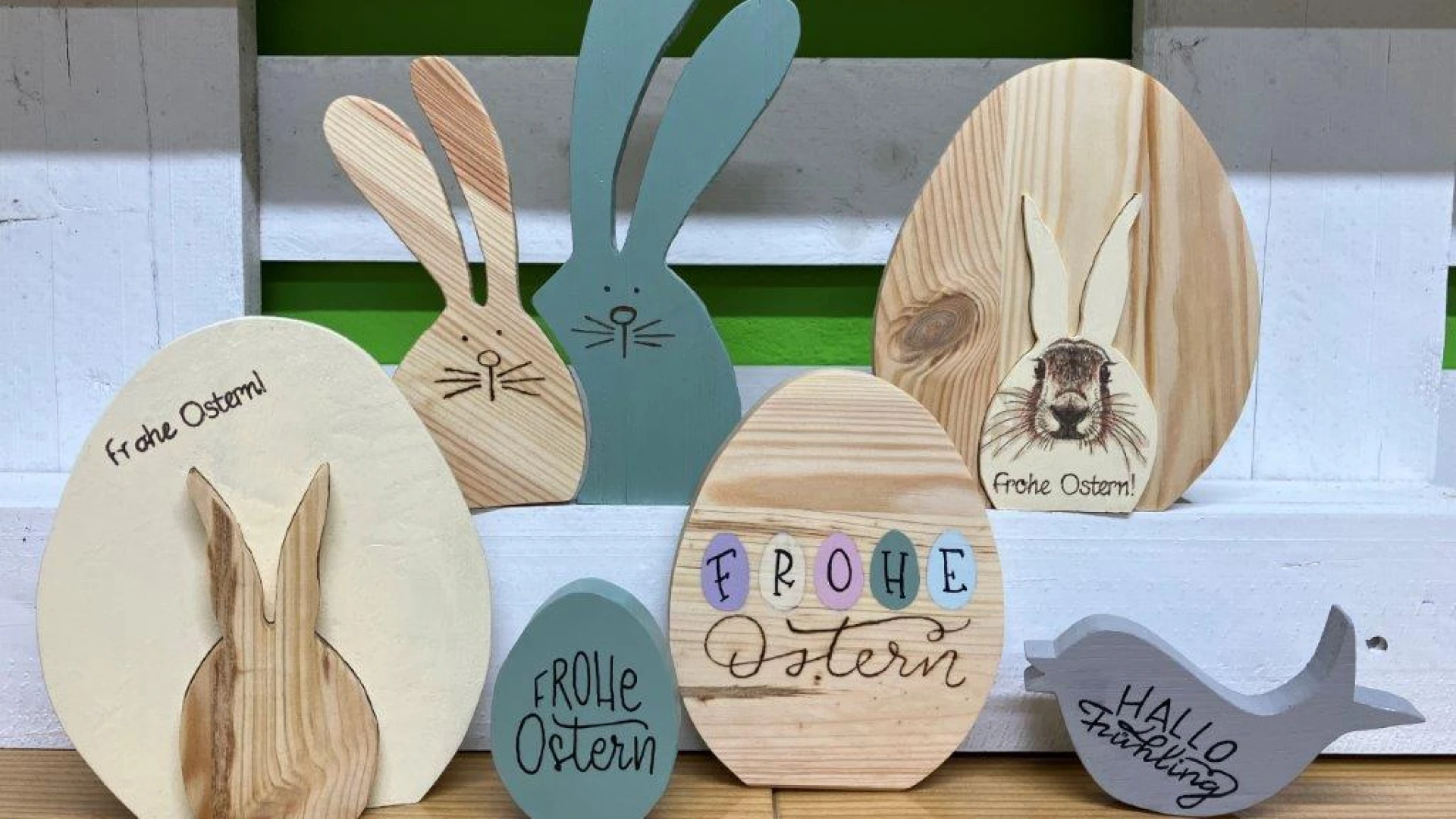 Mehrere Holzfiguren in Form von Osterhasen und Eiern in verschiedenen Farben sind dekorativ auf einem Holzuntergrund angeordnet. Die Figuren sind in unterschiedlichen Gr&ouml;&szlig;en und Formen gestaltet, mit handschriftlichen Oster-Gr&uuml;&szlig;en wie "Frohe Ostern" und "Hallo Fr&uuml;hling" verziert.
