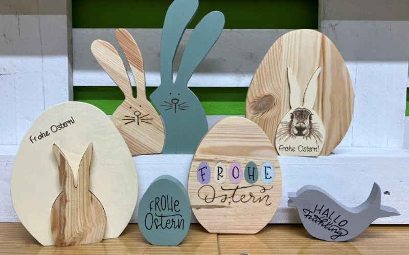 Mehrere Holzfiguren in Form von Osterhasen und Eiern in verschiedenen Farben sind dekorativ auf einem Holzuntergrund angeordnet. Die Figuren sind in unterschiedlichen Gr&ouml;&szlig;en und Formen gestaltet, mit handschriftlichen Oster-Gr&uuml;&szlig;en wie "Frohe Ostern" und "Hallo Fr&uuml;hling" verziert.