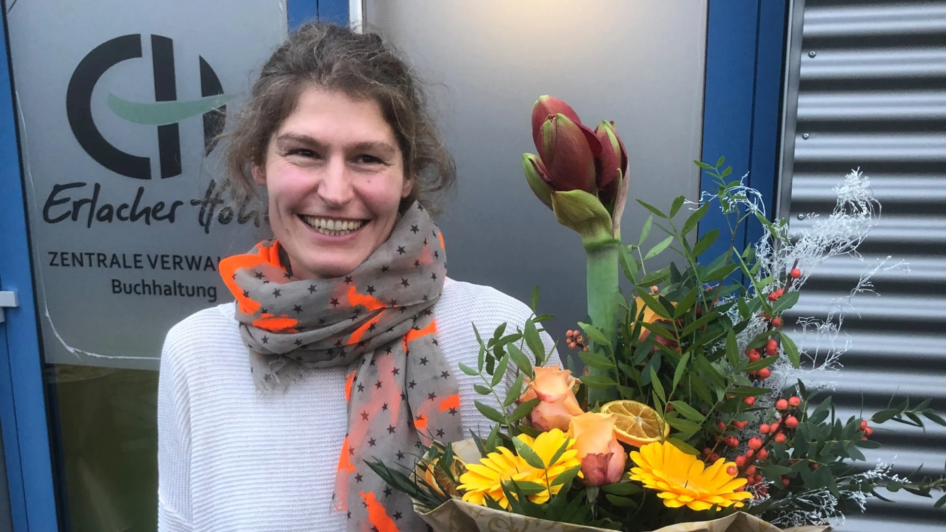 Eine Frau mit dunklem Haar steht l&auml;chelnd vor einem Geb&auml;ude mit einem Blumenstrau&szlig; in der Hand. Sie tr&auml;gt ein wei&szlig;es Oberteil und einen Schal. Im Hintergrund ist eine T&uuml;r zu sehen mit dem Erlacher H&ouml;he Logo und der Aufschrift: "Zentrale Verwaltung Buchhaltung".