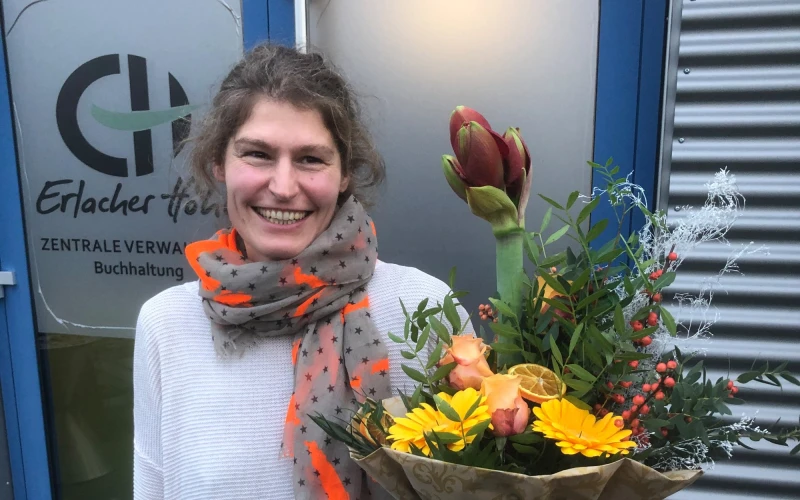 Eine Frau mit dunklem Haar steht l&auml;chelnd vor einem Geb&auml;ude mit einem Blumenstrau&szlig; in der Hand. Sie tr&auml;gt ein wei&szlig;es Oberteil und einen Schal. Im Hintergrund ist eine T&uuml;r zu sehen mit dem Erlacher H&ouml;he Logo und der Aufschrift: "Zentrale Verwaltung Buchhaltung".
