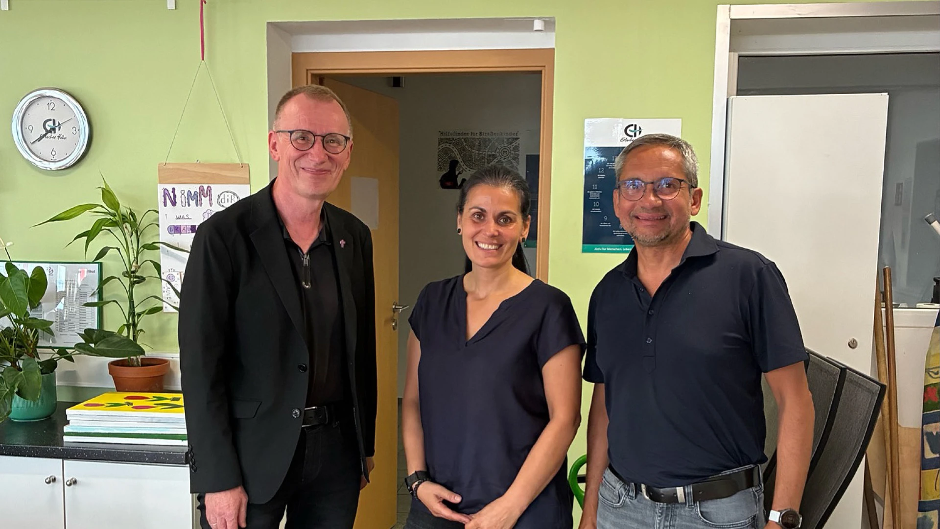 Drei Personen stehen nebeneinander in einem hellen Raum mit gr&uuml;nlichen W&auml;nden