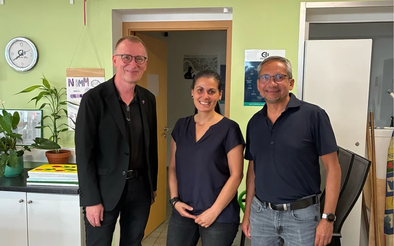 Drei Personen stehen nebeneinander in einem hellen Raum mit gr&uuml;nlichen W&auml;nden