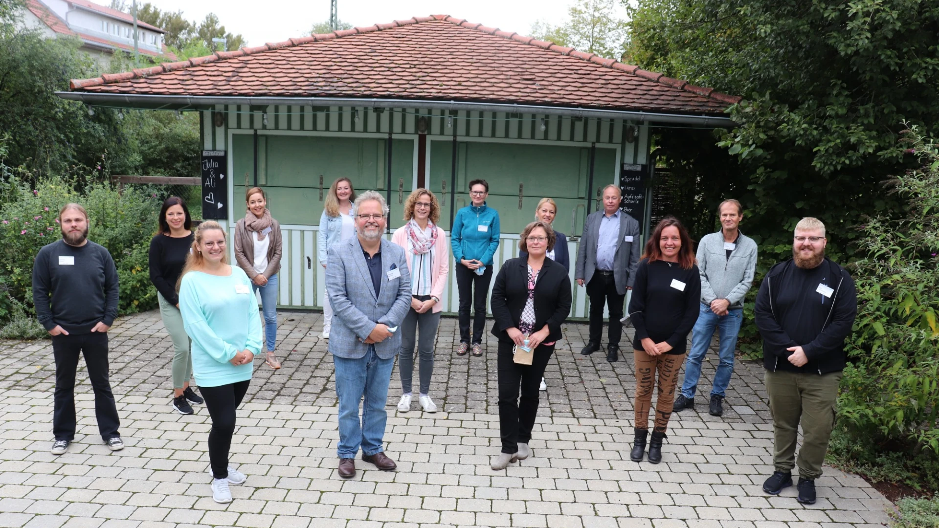 Vierzehn Personen stehen auf einer gepflasterten Fl&auml;che und blicken l&auml;chelnd in die Kamera. Im Hintergrund ist ein kleines Gartengeb&auml;ude zu sehen.
