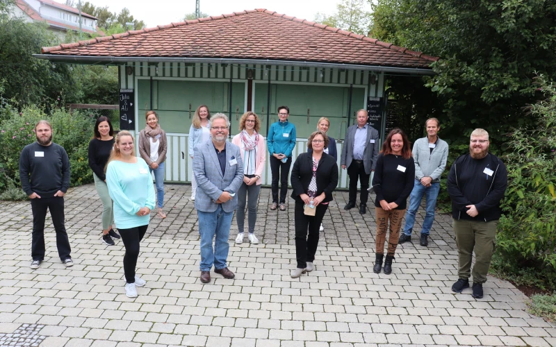 Vierzehn Personen stehen auf einer gepflasterten Fl&auml;che und blicken l&auml;chelnd in die Kamera. Im Hintergrund ist ein kleines Gartengeb&auml;ude zu sehen.