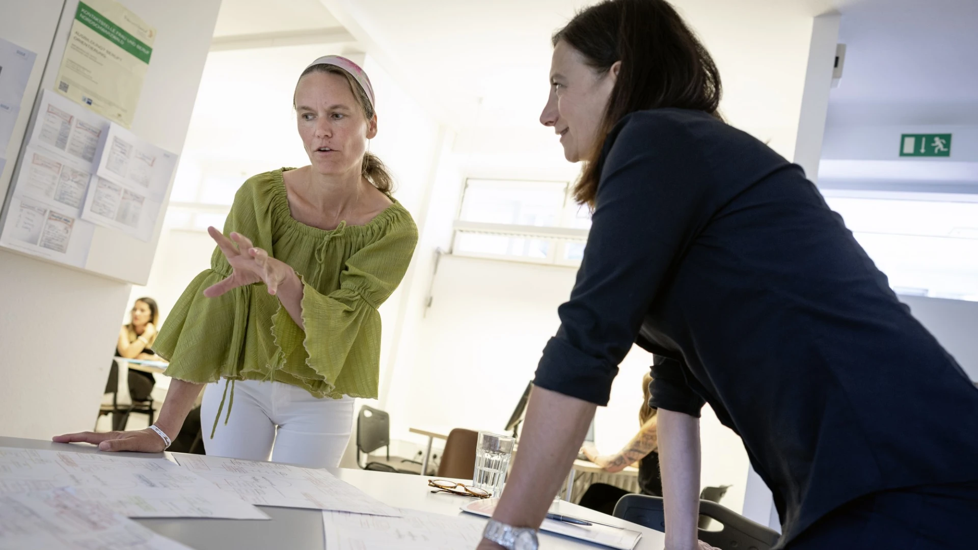 Zwei Frauen lehnen an einem Tisch voller Dokumente. Die Frau links tr&auml;gt eine olivgr&uuml;ne Bluse und wei&szlig;e Hose, die andere tr&auml;gt schwarzer Kleidung. Im Hintergrund sind weitere Tische und Personen zu sehen.