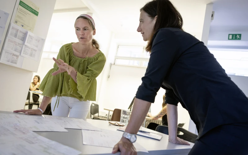 Zwei Frauen lehnen an einem Tisch voller Dokumente. Die Frau links tr&auml;gt eine olivgr&uuml;ne Bluse und wei&szlig;e Hose, die andere tr&auml;gt schwarzer Kleidung. Im Hintergrund sind weitere Tische und Personen zu sehen.