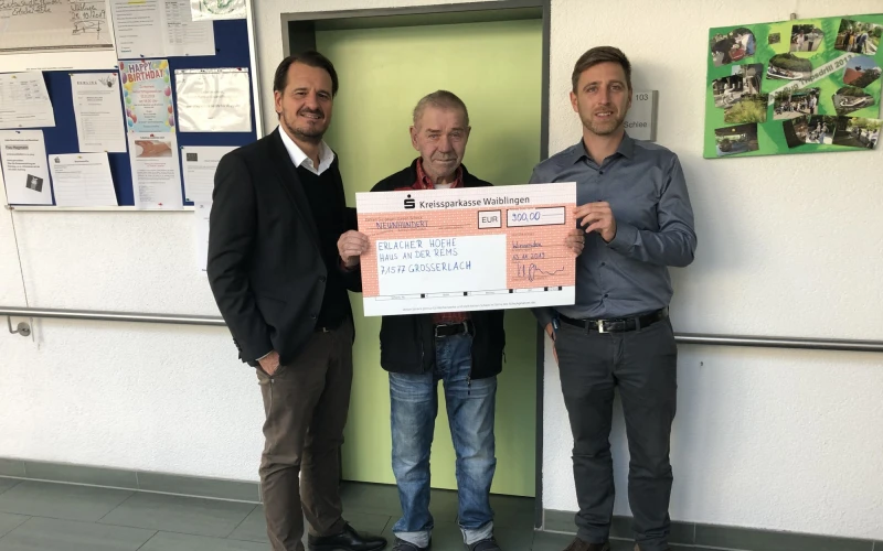 Drei M&auml;nner stehen in einem Geb&auml;ude vor einer hellgr&uuml;nen T&uuml;re. Der mittlere Mann h&auml;lt gemeinsam mit der rechten Person einen Scheck in der Hand. Im Hintergrund ist eine Pinnwand mit Zetteln und ein Plakat an der Wand zu sehen.