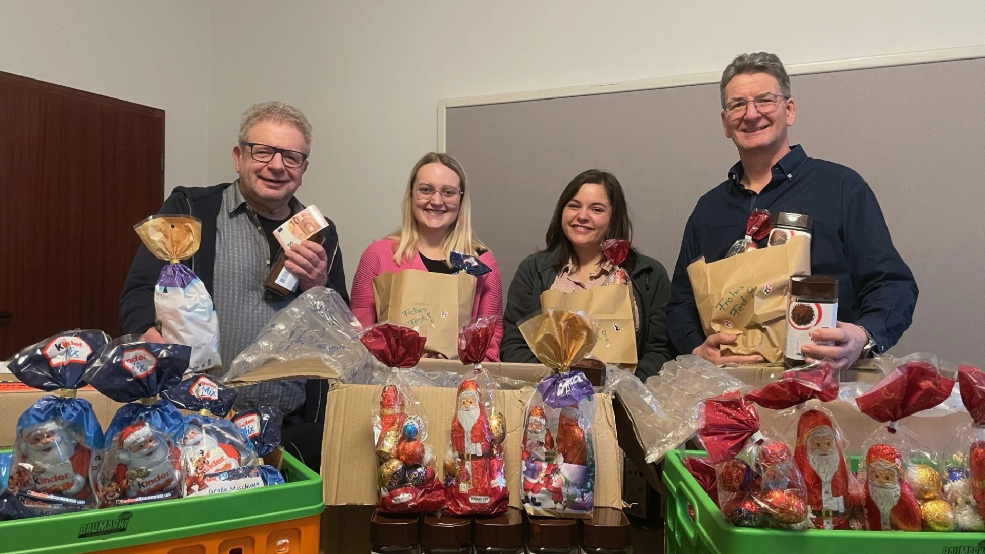 Vier Personen l&auml;cheln in die Kamera. Sie stehen hinter Kisten und Kartons mit Weihnachtsgeschenken.