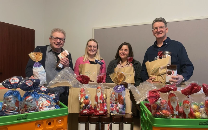 Vier Personen l&auml;cheln in die Kamera. Sie stehen hinter Kisten und Kartons mit Weihnachtsgeschenken.