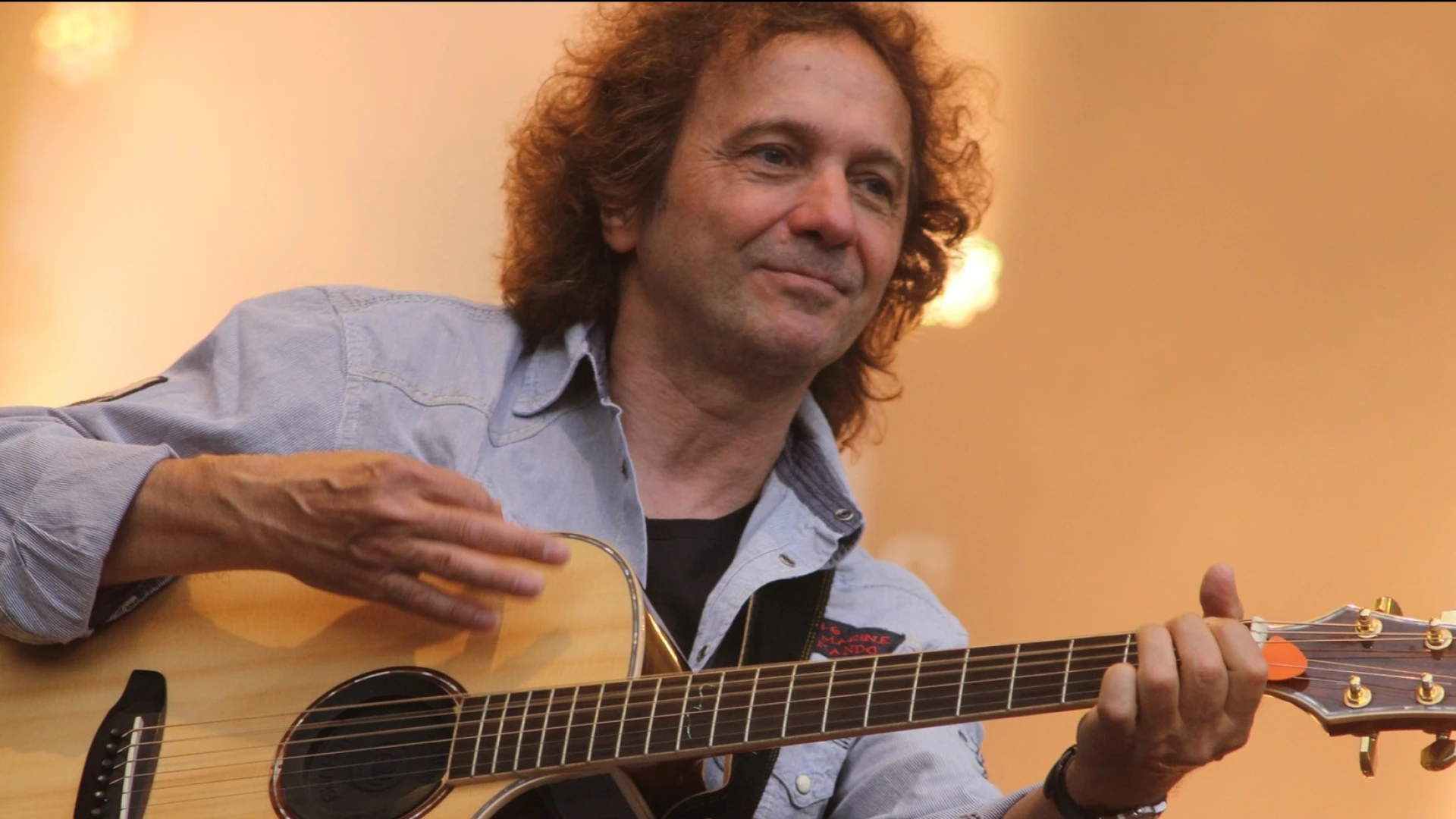 Ein Mann mit langen, welligen Haaren h&auml;lt eine Gitarre in der Hand und tr&auml;gt ein blaues Hemd. Im Hintergrund sind Scheinwerferlichter zu sehen.