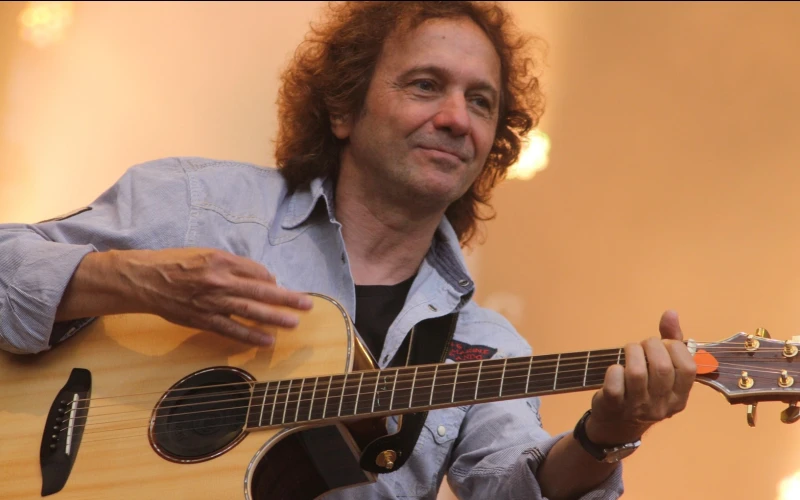 Ein Mann mit langen, welligen Haaren h&auml;lt eine Gitarre in der Hand und tr&auml;gt ein blaues Hemd. Im Hintergrund sind Scheinwerferlichter zu sehen.