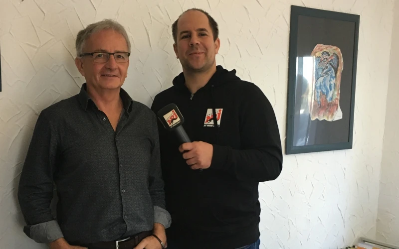 Zwei M&auml;nner stehen vor einer wei&szlig;en Wand. Der Mann rechts h&auml;lt in seiner linken Hand einen Mikrofon. Im Hintergrund ist ein gerahmtes Bild an der Wand zu sehen.