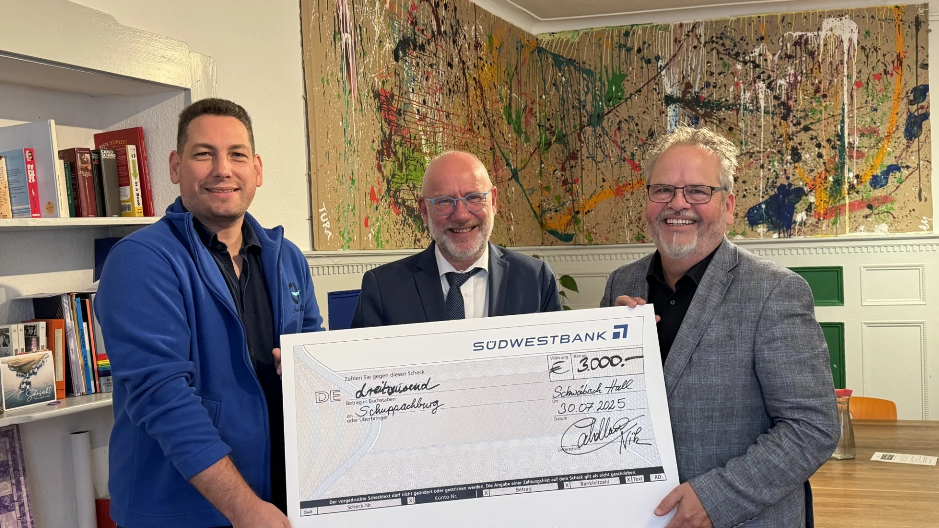 Drei M&auml;nner halten gemeinsam einen gro&szlig;en Scheck mit der Aufschrift "S&uuml;dwestbank" in einem B&uuml;ro
