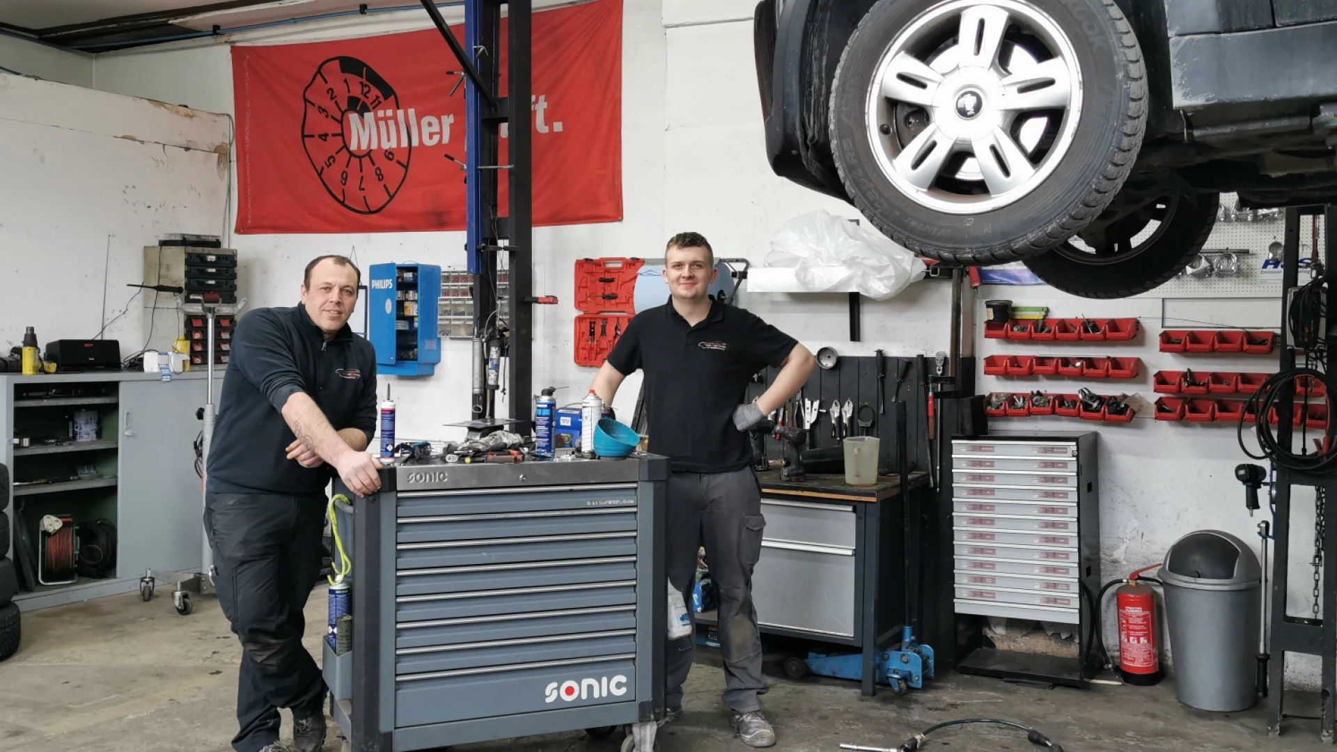Zwei Personen stehen in einer Autowerkstatt neben einer Hebeb&uuml;hne, auf der ein Fahrzeug ist. Beide lehnen sich an einen Werkstattwagen. Im Hintergrund sind weitere Werkzeuge, Schr&auml;nke, Regale und Autoreifen zu sehen.