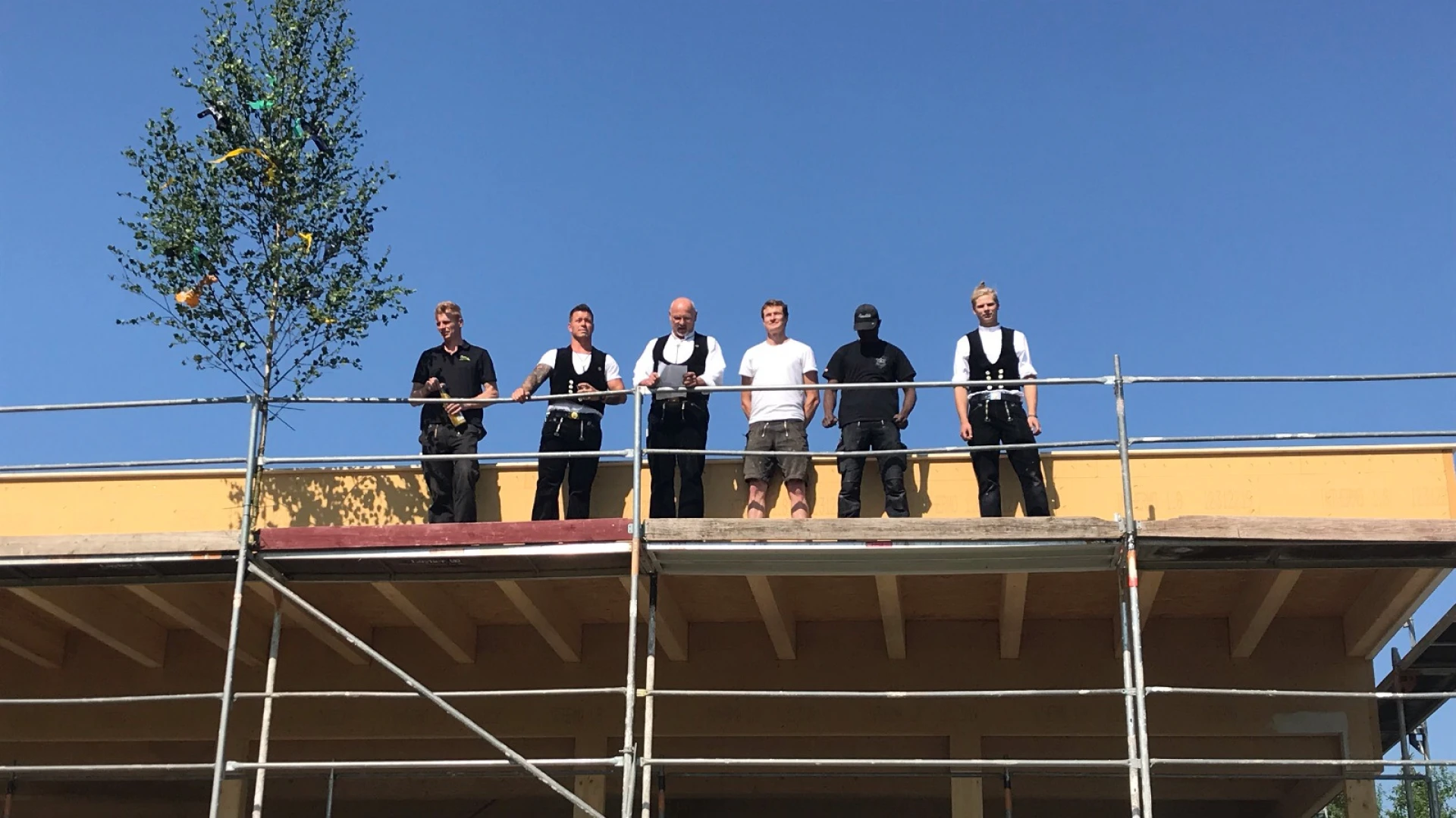 Sechs Personen stehen auf einem Ger&uuml;st vor einem Dachvorsprung. Die M&auml;nner sind in schwarezn Hosen gekleidet, manche tragen ein wei&szlig;es T-Shirt und eine schwarze Weste. Es scheint ein Richtfest gefeiert zu werden.