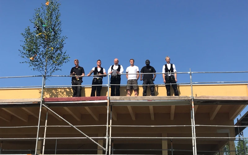 Sechs Personen stehen auf einem Ger&uuml;st vor einem Dachvorsprung. Die M&auml;nner sind in schwarezn Hosen gekleidet, manche tragen ein wei&szlig;es T-Shirt und eine schwarze Weste. Es scheint ein Richtfest gefeiert zu werden.