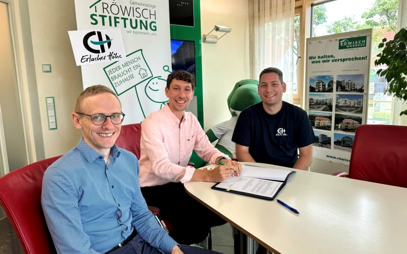 Drei M&auml;nner sitzen an einem Tisch mit Dokumenten. Es scheint sich um die Unterzeichnung eines Vertrages zu handeln. Alle drei M&auml;nner blicken freudig l&auml;chelnd in die Kamera.