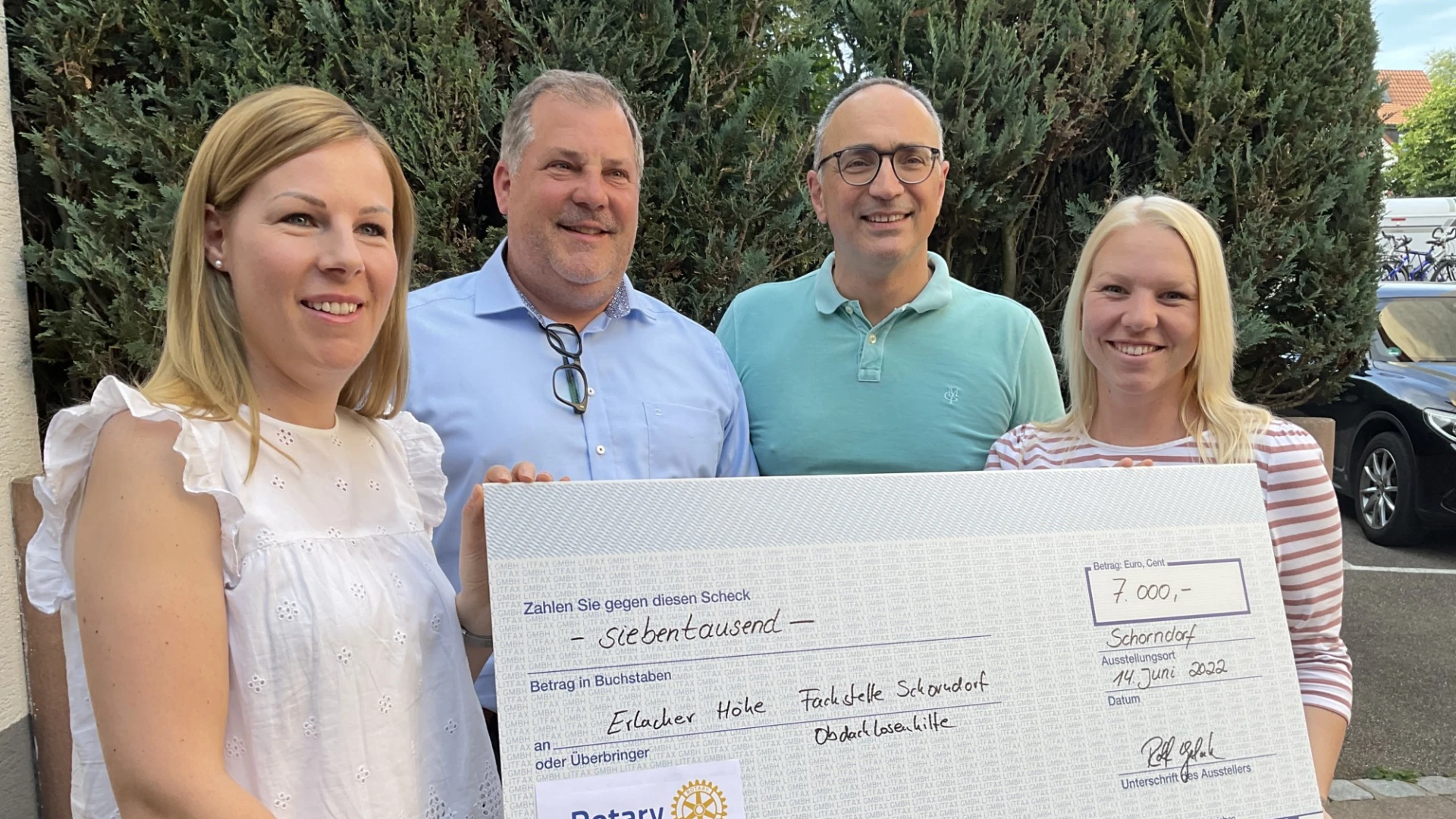 Zwei M&auml;nner stehen zwischen zwei Frauen, die einen Spendenscheck in der Hand halten. Alle l&auml;cheln in die Kamera. Im Hintergrund sind eine Hecke und parkende Autos zu sehen.