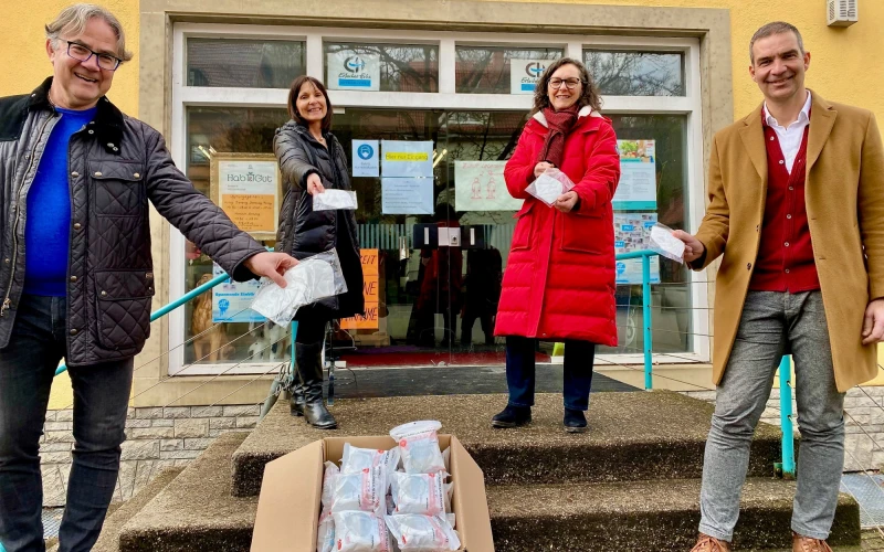 Vier Personen stehen auf einer Treppe vor einem Geb&auml;ude und halten FFP3-MAsen in den H&auml;nden. Vor ihnen steht ein ge&ouml;ffneter Karton mit weiteren FFP3-Masken. Im Fenster des Geb&auml;udes im Hintergrund sind Plakate und Informationen ausgeh&auml;ngt.
