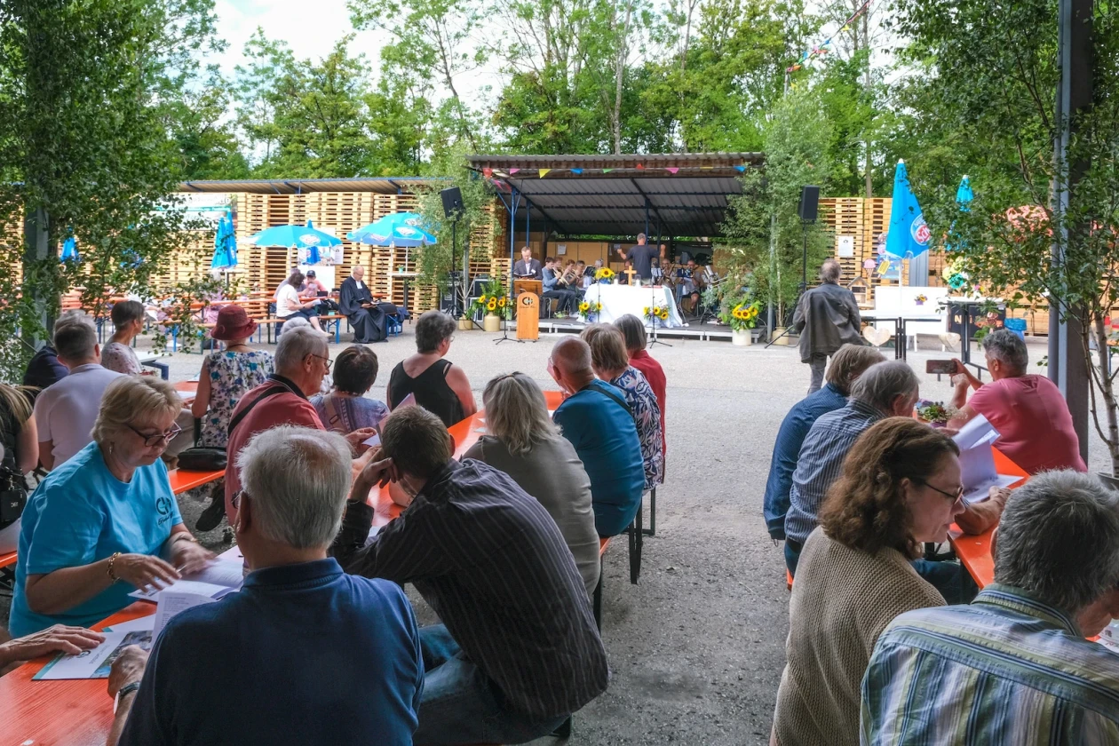 Ein Gel&auml;nde im Gr&uuml;nen mit vielen Menschen, die auf B&auml;nken sitzen. Auf einer B&uuml;hne steht ein Mann an einem Pult und spricht.