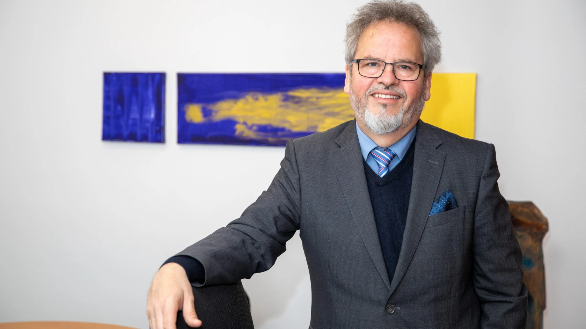 L&auml;chelnder &auml;lterer Mann im grauen Anzug, hellblauem Hemd unter blauem Pullover und gestreifter Krawatte, der in einem B&uuml;ro vor Kunstwerken in Blau und Gelb steht. Er tr&auml;gt eine Brille und hat graues, welliges Haar und einen grauen Bart.