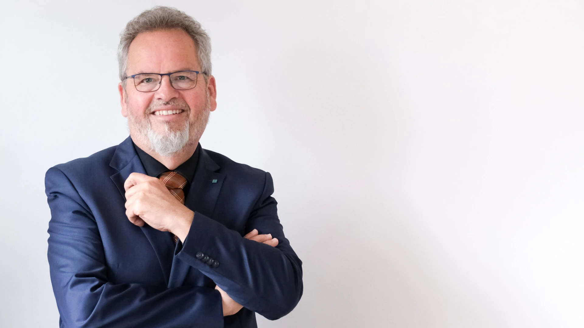 Ein Mann mit grauem Haar und Brille tr&auml;gt ein dunkelblaues Sakko und l&auml;chelt freundlich in die Kamera. Er steht vor einem wei&szlig;en Hintergrund mit verschr&auml;nkten Armen in entspannter Pose.