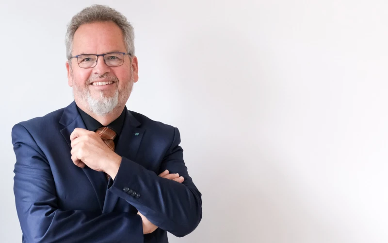 Ein Mann mit grauem Haar und Brille tr&auml;gt ein dunkelblaues Sakko und l&auml;chelt freundlich in die Kamera. Er steht vor einem wei&szlig;en Hintergrund mit verschr&auml;nkten Armen in entspannter Pose.