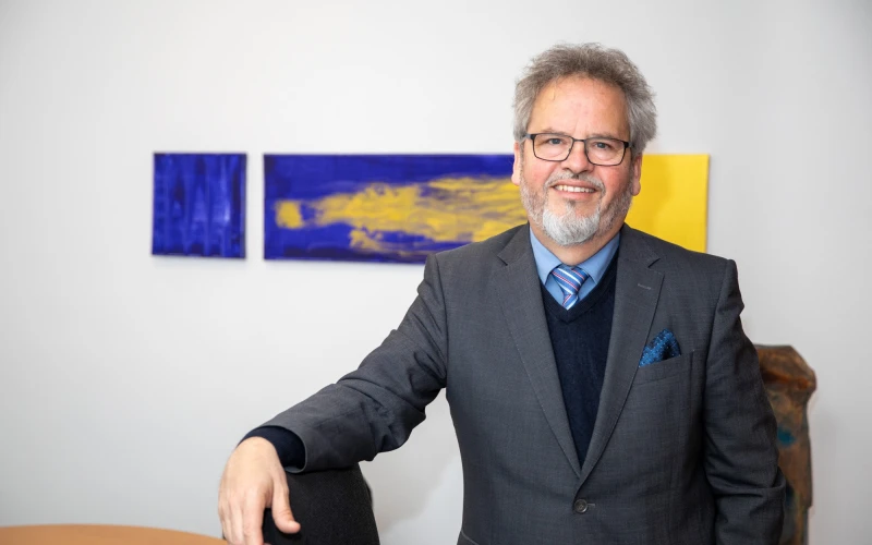 L&auml;chelnder &auml;lterer Mann im grauen Anzug, hellblauem Hemd unter blauem Pullover und gestreifter Krawatte, der in einem B&uuml;ro vor Kunstwerken in Blau und Gelb steht. Er tr&auml;gt eine Brille und hat graues, welliges Haar und einen grauen Bart.