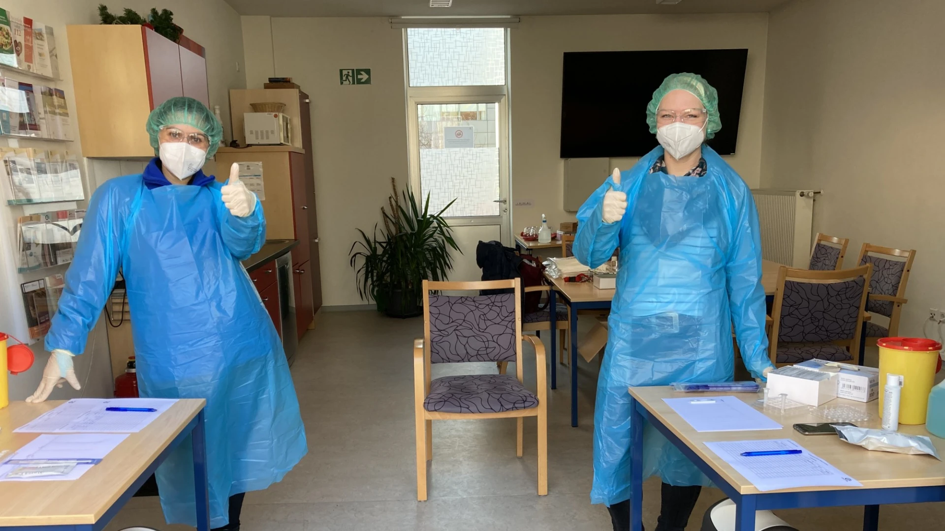 Zwei Personen in blauer Schutzkleidung stehen in einem Raum und zeigen mit dem Daumen nach oben. Vor ihnen stehen Tische mit Formularen, Testmaterialien und Desinfektionsmittel. Im Hintergrund sind St&uuml;hle, ein Schrank und ein gro&szlig;es Fenster zu sehen.