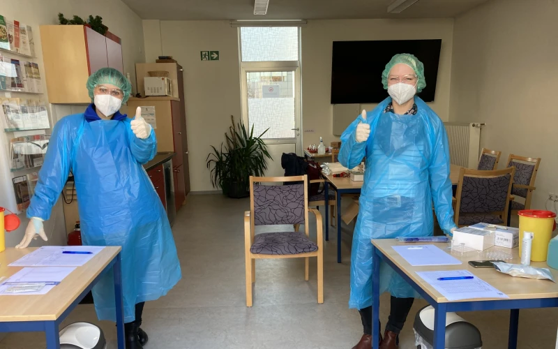 Zwei Personen in blauer Schutzkleidung stehen in einem Raum und zeigen mit dem Daumen nach oben. Vor ihnen stehen Tische mit Formularen, Testmaterialien und Desinfektionsmittel. Im Hintergrund sind St&uuml;hle, ein Schrank und ein gro&szlig;es Fenster zu sehen.