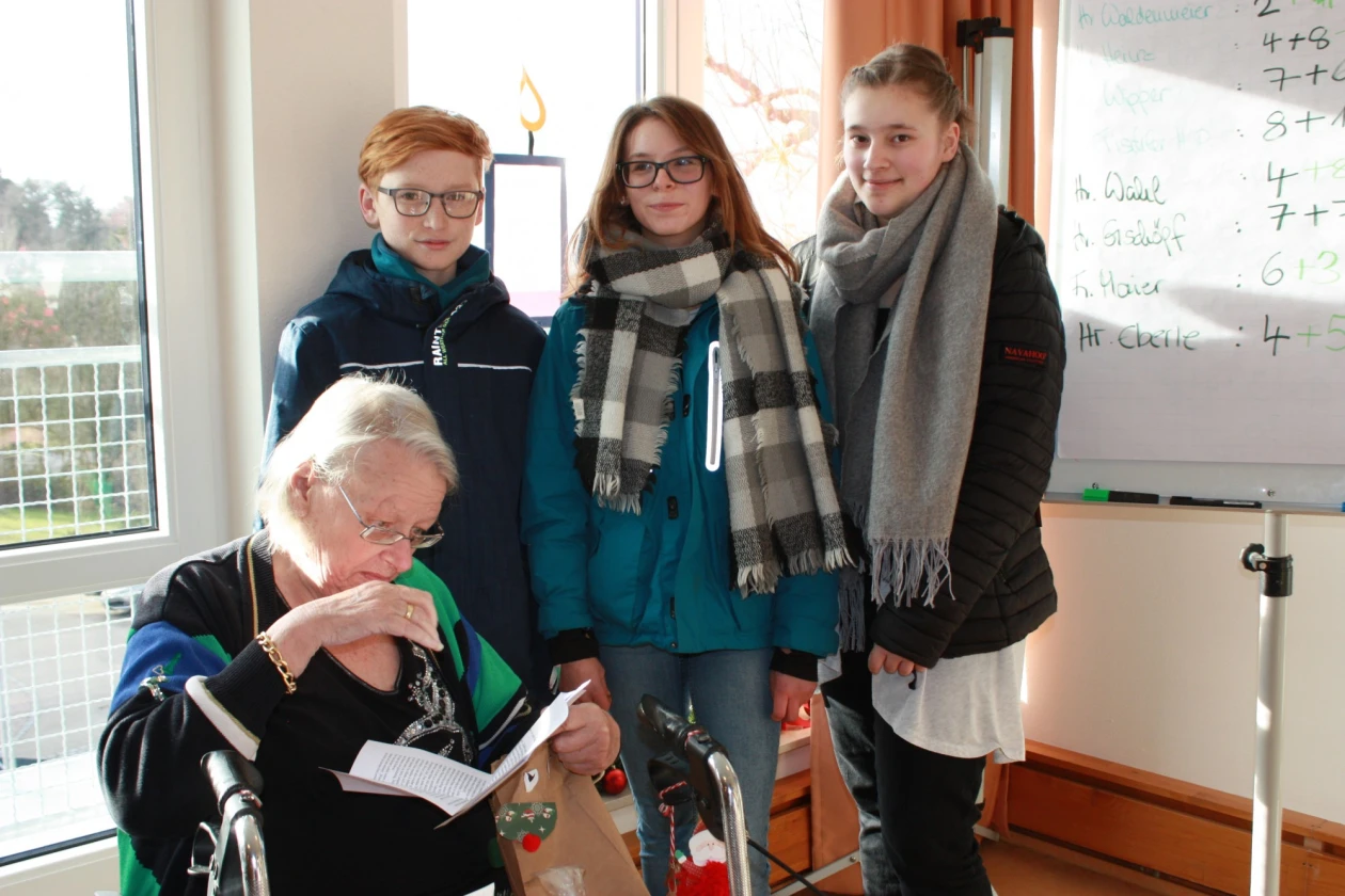 Eine Frau im Rollstuhl liest einen Brief. Neben ihr stehen drei Jugendliche in warmer Winterkleidung. Im Raum befinden sich ein Whiteboard und ein Fenster.