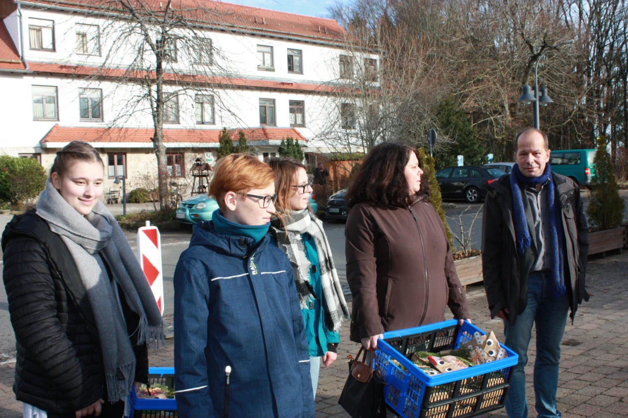 F&uuml;nf Personen stehen vor einem wei&szlig;en Geb&auml;ude auf einer gepflasterten Fl&auml;che. Sie tragen zwei Klappboxen, die mit verpackten Geschenken in braunen Papiertaschen mit aufgeklebten Wackelaugen liegen. Im Hintergrund sind geparkte Autos und ein Geb&auml;ude zu erkennen.