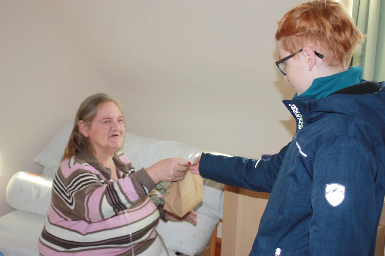 Ein Kind mit r&ouml;tlichen Haaren in blauer Jacke &uuml;berreicht einer &auml;lteren Frau mit gestreiftem Oberteil eine Papiert&uuml;te. Die Frau sitzt auf einem Bett.
