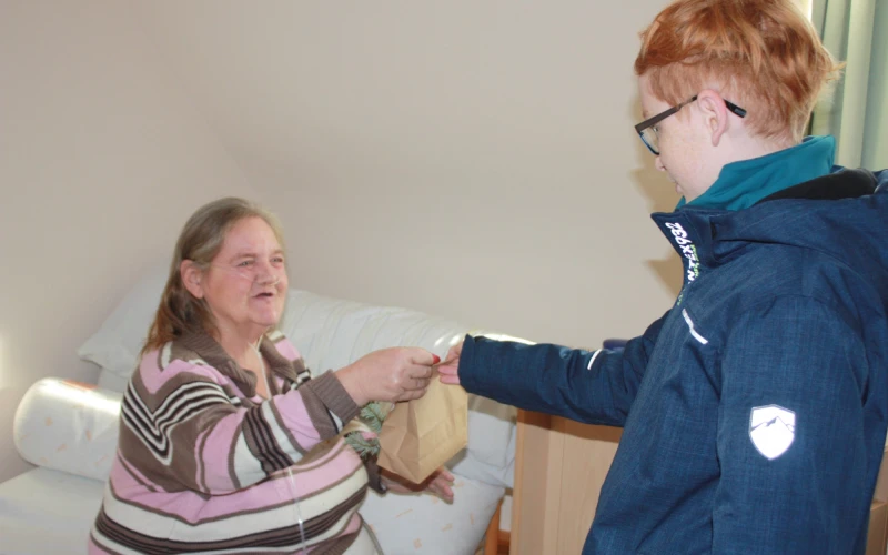 Ein Kind mit r&ouml;tlichen Haaren in blauer Jacke &uuml;berreicht einer &auml;lteren Frau mit gestreiftem Oberteil eine Papiert&uuml;te. Die Frau sitzt auf einem Bett.