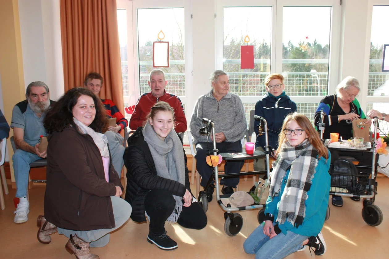 Eine Gruppe von Menschen unterschiedlichen Alters befindet sich in einem hellen Raum mit gro&szlig;en Fenstern. Ein orangefarbiger Vorhang und zwei Rollatoren sind im Raum zu sehen.
