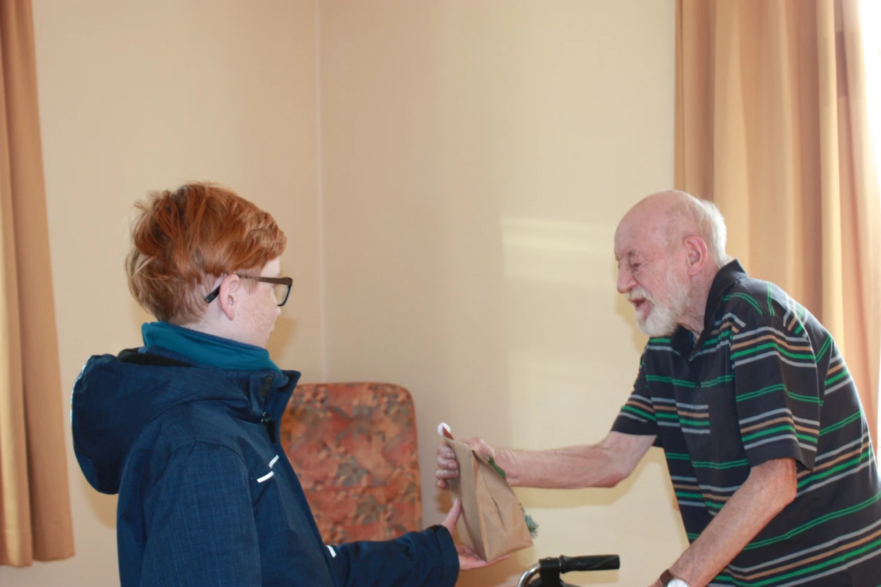 Ein &auml;lterer Mann mit wei&szlig;em Bart und ein Kind mit r&ouml;tlichen Haaren ist zu sehen. Beide halten gemeinsam eine Papiertasche in der Hand. Das Kind tr&auml;gt eine blaue Jacke. Im Hintergrund ist ein gebl&uuml;mter Sessel, sowie Vorh&auml;nge an den Fenstern zu sehen.