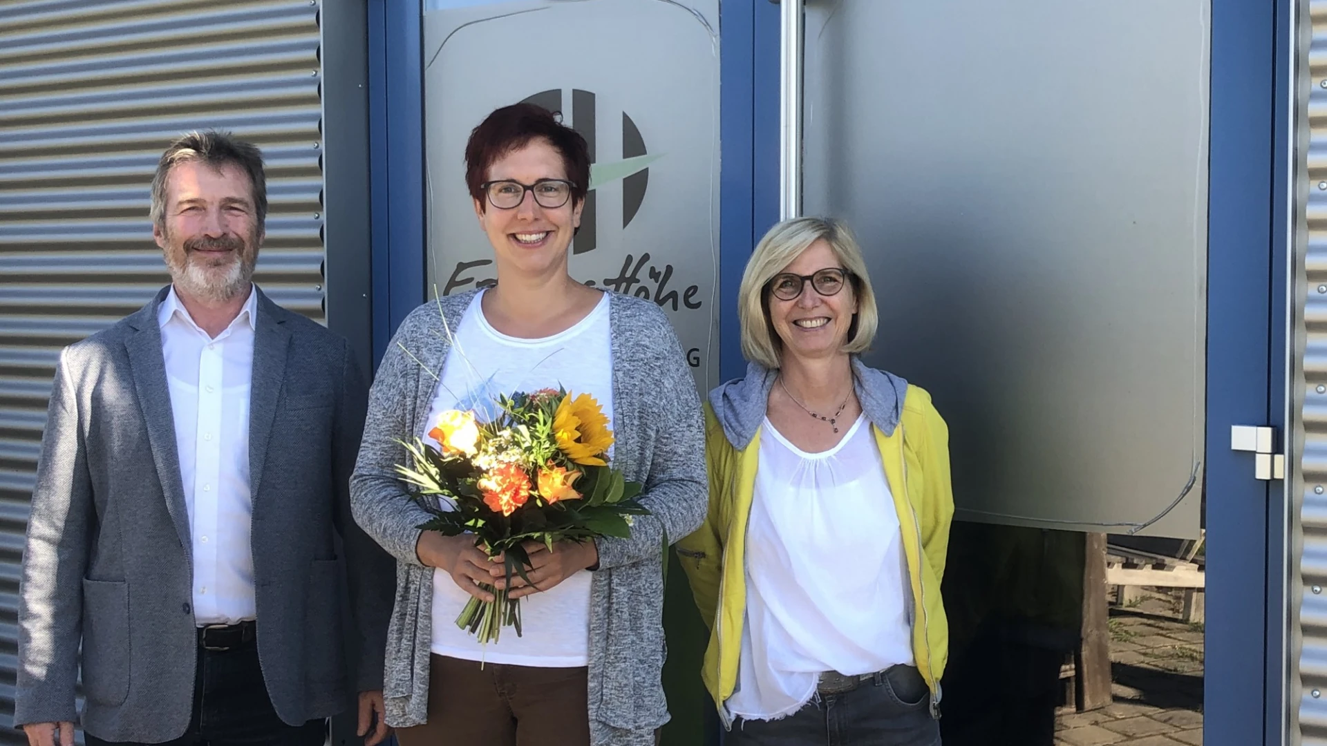 Drei Personen stehen vor einer T&uuml;r, die mit dem Logo der Erlacher H&ouml;he versehen ist. Die Frau in der Mitte h&auml;lt einen Blumenstrau&szlig; in den H&auml;nden. Alle drei l&auml;cheln in die Kamera.